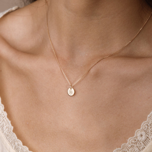A delicate gold chain necklace with a small circular disc pendant featuring an engraved sun design, shown worn on a model's neck.