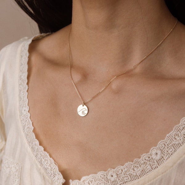 A close-up of a model wearing a delicate gold chain necklace with a small circular pendant engraved with a wave design, styled with a cream lace-trimmed top.