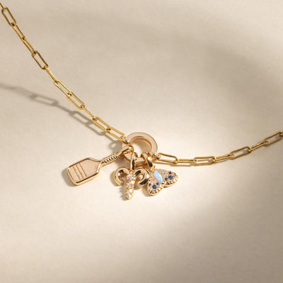 A gold charm necklace with a gold pickleball racket charm, an Aries zodiac symbol charm with clear accent stones, and a moth charm featuring multiple stones in different shades of blue, set against a warm neutral back drop.