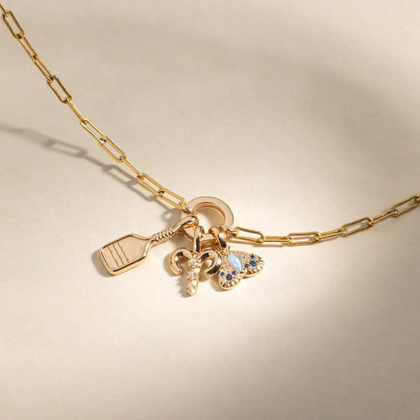 A gold charm necklace with a gold pickleball racket charm, an Aries zodiac symbol charm with clear accent stones, and a moth charm featuring multiple stones in different shades of blue, set against a warm neutral back drop.