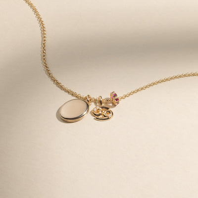 A gold chain necklace with a gold oval locket pendant, a Cancer zodiac symbol charm with accent stones, and a round gold charm with square red gemstones.