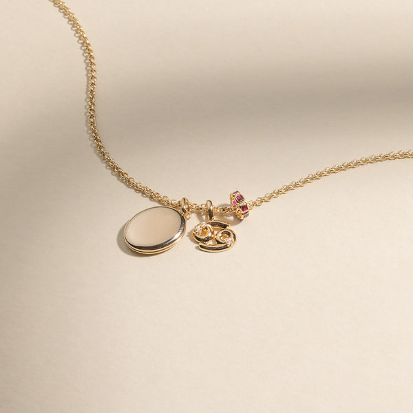 A gold chain necklace with a gold oval locket pendant, a Cancer zodiac symbol charm with accent stones, and a round gold charm with square red gemstones.