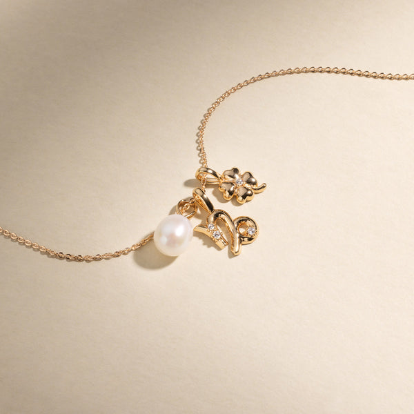 A gold chain necklace with a round pearl charm, a Capricorn zodiac symbol charm with clear accent stones, and a four leaf clover charm with one clear stone in the center.