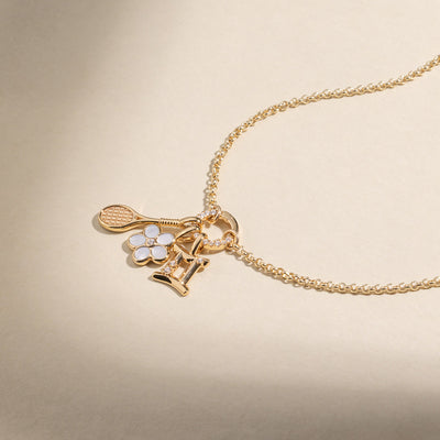 A gold chain necklace with a gold tennis racket charm, a gold daisy charm with white stones and clear center stone, and a Gemini Zodiac charm with clear accent stones, set against a warm beige background.