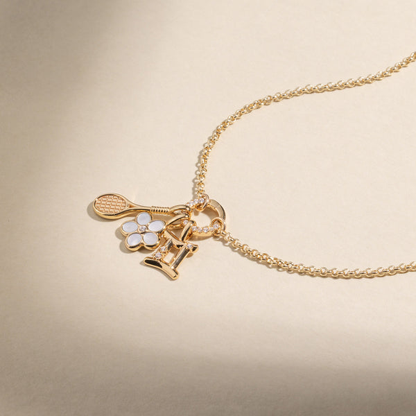 A gold chain necklace with a gold tennis racket charm, a gold daisy charm with white stones and clear center stone, and a Gemini Zodiac charm with clear accent stones, set against a warm beige background.