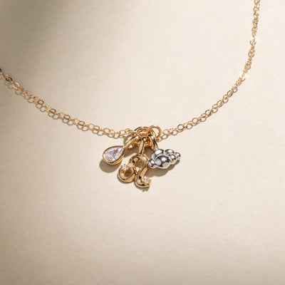 A gold chain necklace featuring a gold pear bezel-set charm, a Leo zodiac symbol charm with clear accent stones, and a silver cloud charm with a gold jump ring, set against a neutral beige background.