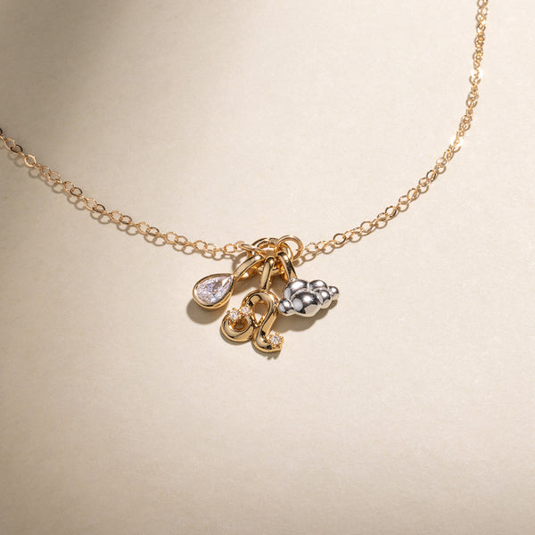 A gold chain necklace featuring a gold pear bezel-set charm, a Leo zodiac symbol charm with clear accent stones, and a silver cloud charm with a gold jump ring, set against a neutral beige background.