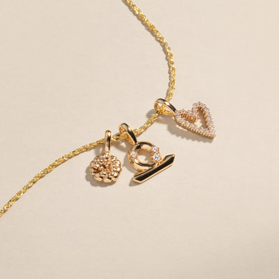 A gold delicate chain necklace with a marigold charm, a Libra zodiac symbol charm with clear accent stones, and a heart outline charm covered in clear stones.