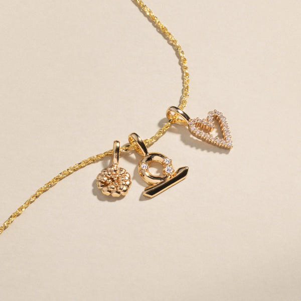 A gold delicate chain necklace with a marigold charm, a Libra zodiac symbol charm with clear accent stones, and a heart outline charm covered in clear stones.