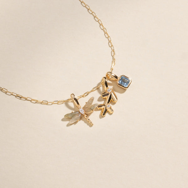 A gold chain necklace with a dragonfly charm with natural gemstone wings and a clear stone center, a Sagittarius zodiac symbol charm with clear accent stones, and a square light blue gemstone charm.