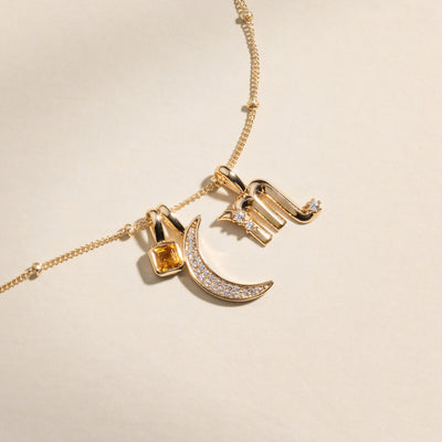 A gold dainty chain necklace with a square, amber colored gemstone charm, a crescent moon charm with clear stones, and a Scorpio zodiac symbol charm with clear accent stones.