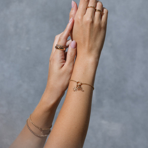 Close-up of hands wearing multiple gold rings, two gold chain bracelets, and gold charm bracelet featuring various charms, set against a blue-grey backdrop.