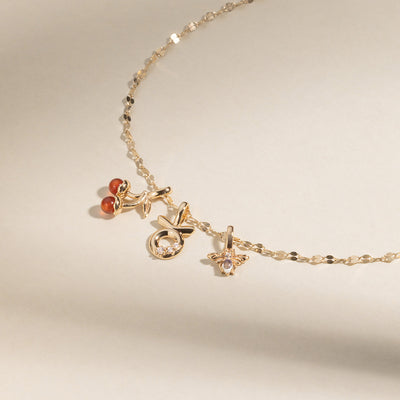 A gold chain necklace with a gold cherry charm with two red agate stones, a Taurus zodiac symbol charm with clear accent stones, and a bee charm with two clear stones, set against a neutral background.