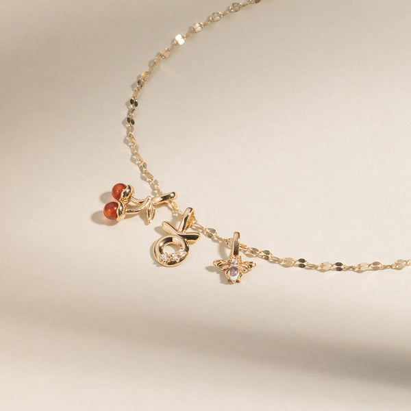 A gold chain necklace with a gold cherry charm with two red agate stones, a Taurus zodiac symbol charm with clear accent stones, and a bee charm with two clear stones, set against a neutral background.