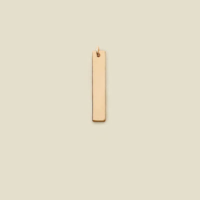 A polished gold vertical rectangle bar charm, shown on a solid light beige background.