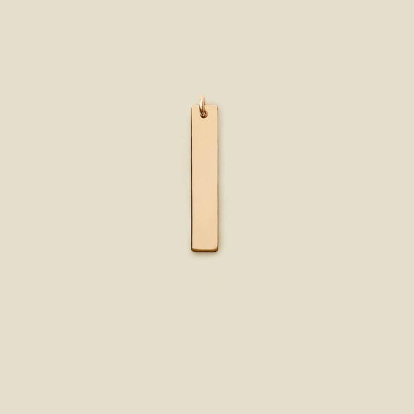 A polished gold vertical rectangle bar charm, shown on a solid light beige background.