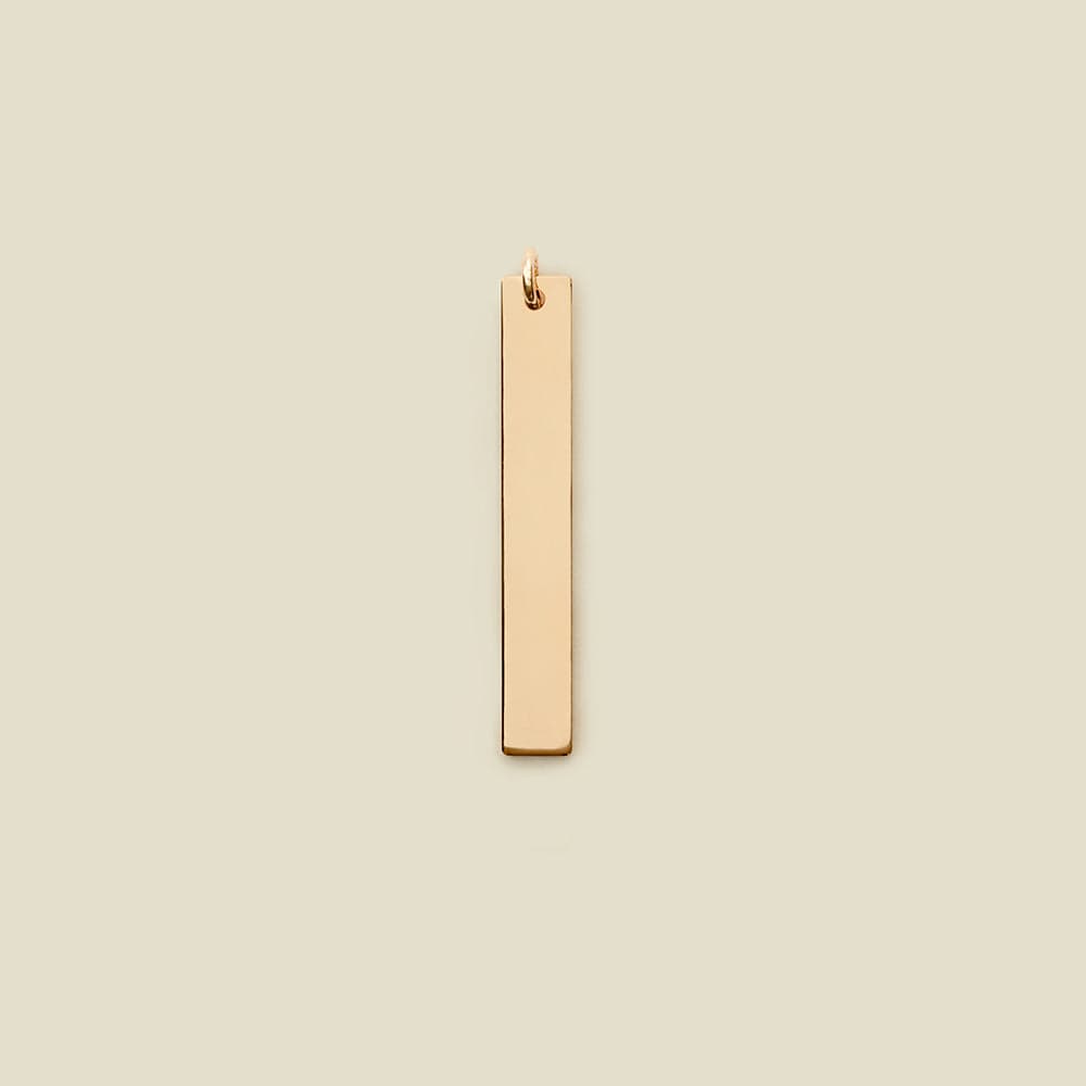 A polished gold vertical rectangle bar charm, shown on a solid light beige background.