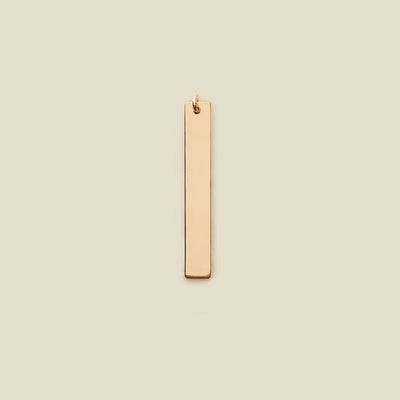 A polished gold vertical rectangle bar charm, shown on a solid light beige background.