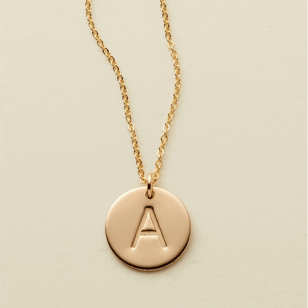A gif of a delicate gold pendant chain with flat round disc pendants, each engraved with a single initial. The gif starts with a single disc, then continues adding one after another up to three discs.
