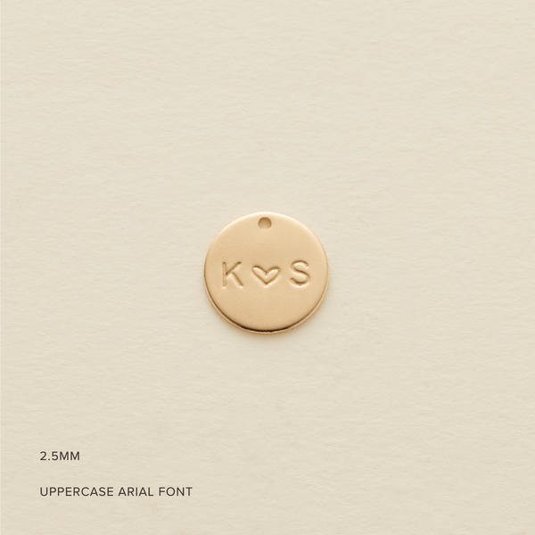 A round flat gold disc pendant engraved with the letters 'K' and 'S' separated by a heart, shown on a textured light beige background. In the bottom left corner are the words '2.5MM' and 'UPPERCASE ARIAL FONT'.