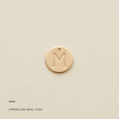 A round flat gold disc pendant with an engraved initial 'M', shown on a textured light beige background. In the bottom left corner are the words '6MM' and 'UPPERCASE ARIAL FONT'.