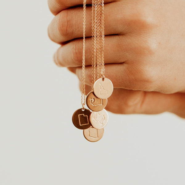 A person holding five delicate gold pendant chain necklaces, each with a single flat round disc engraved with the outline of a state.