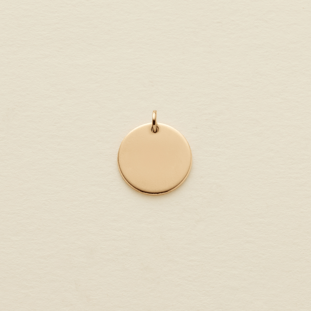 A small, flat, circular gold pendant with a top loop, centered on a textured off-white background.