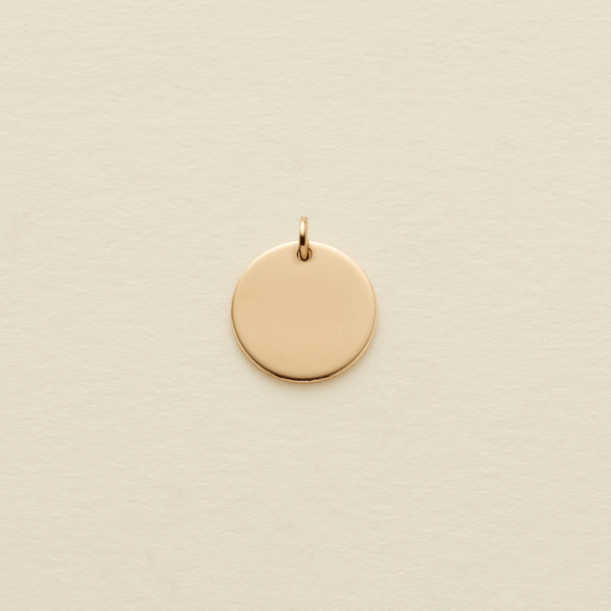 A small, flat, circular gold pendant with a top loop, centered on a textured off-white background.