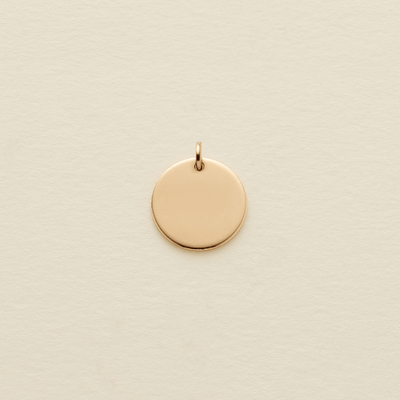 A small, flat, circular gold pendant with a top loop, centered on a textured off-white background.