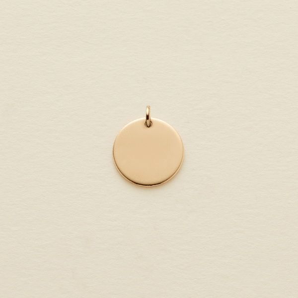 A small, flat, circular gold pendant with a top loop, centered on a textured off-white background.