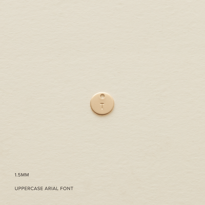 A small flat round gold disc with an uppercase initial 'T' engraved on the front, shown on a textured light beige surface. In the bottom left corner there is text saying '1.5MM' and 'UPPERCASE ARIAL FONT'.