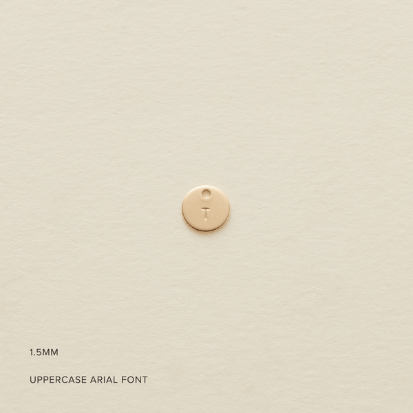 A small flat round gold disc with an uppercase initial 'T' engraved on the front, shown on a textured light beige surface. In the bottom left corner there is text saying '1.5MM' and 'UPPERCASE ARIAL FONT'.
