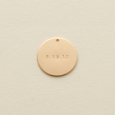 A flat gold round disc pendant with the date '8.15.12' engraved across the center, shown on a textured light beige surface.