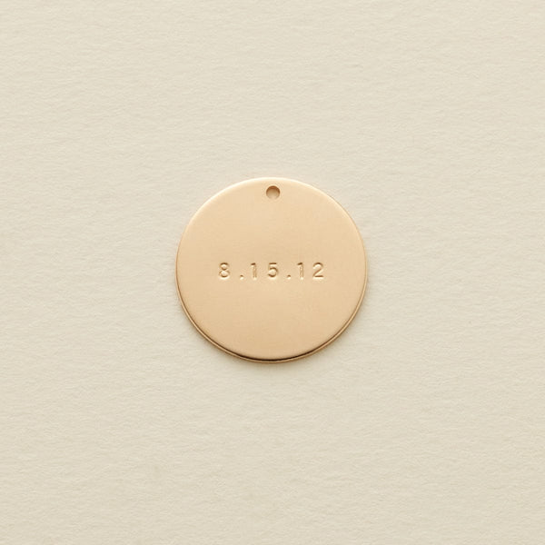 A flat gold round disc pendant with the date '8.15.12' engraved across the center, shown on a textured light beige surface.