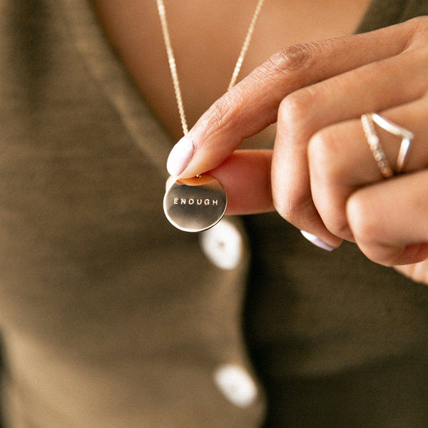 A person holding the delicate gold pendant necklace they are wearing with a round flat disc, engraved with the word 'ENOUGH'.