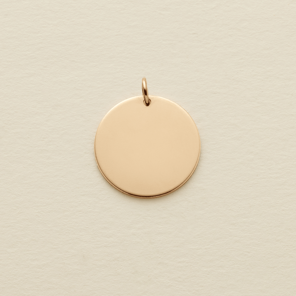 A round flat blank gold pendant with a gold jump ring on the top, shown on a textured light beige surface.