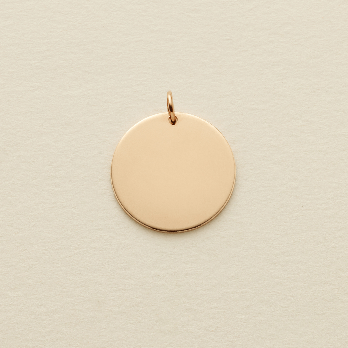 A round flat blank gold pendant with a gold jump ring on the top, shown on a textured light beige surface.