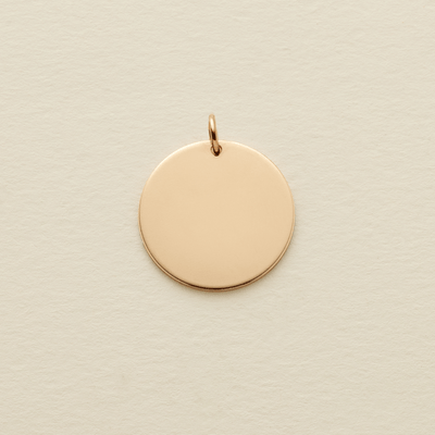 A round flat blank gold pendant with a gold jump ring on the top, shown on a textured light beige surface.