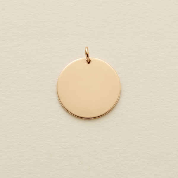 A round flat blank gold pendant with a gold jump ring on the top, shown on a textured light beige surface.