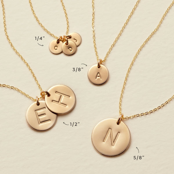 Four different delicate pendant chain necklaces laying flat on a solid light beige surface. Each of the necklaces has a different size and different amount of flat round disc pendants in the sizes '1/4"', '3/8"', '1/2"', and '5/8"'.