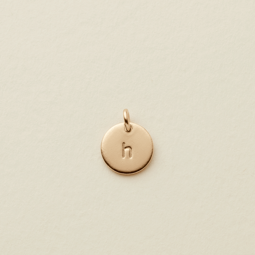 A small round gold disc charm engraved with the lowercase letter 'h' and suspended from a loop, centered on a light cream textured background.