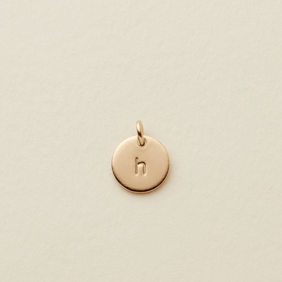 A small round gold disc charm engraved with the lowercase letter 'h' and suspended from a loop, centered on a light cream textured background.