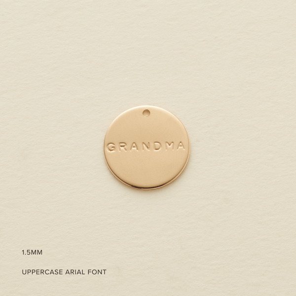 A round flat gold disc pendant with the word 'GRANDMA' engraved, shown on a textured light beige surface. In the bottom corner are the words '1.5MM' and 'UPPERCASE ARIAL FONT'.