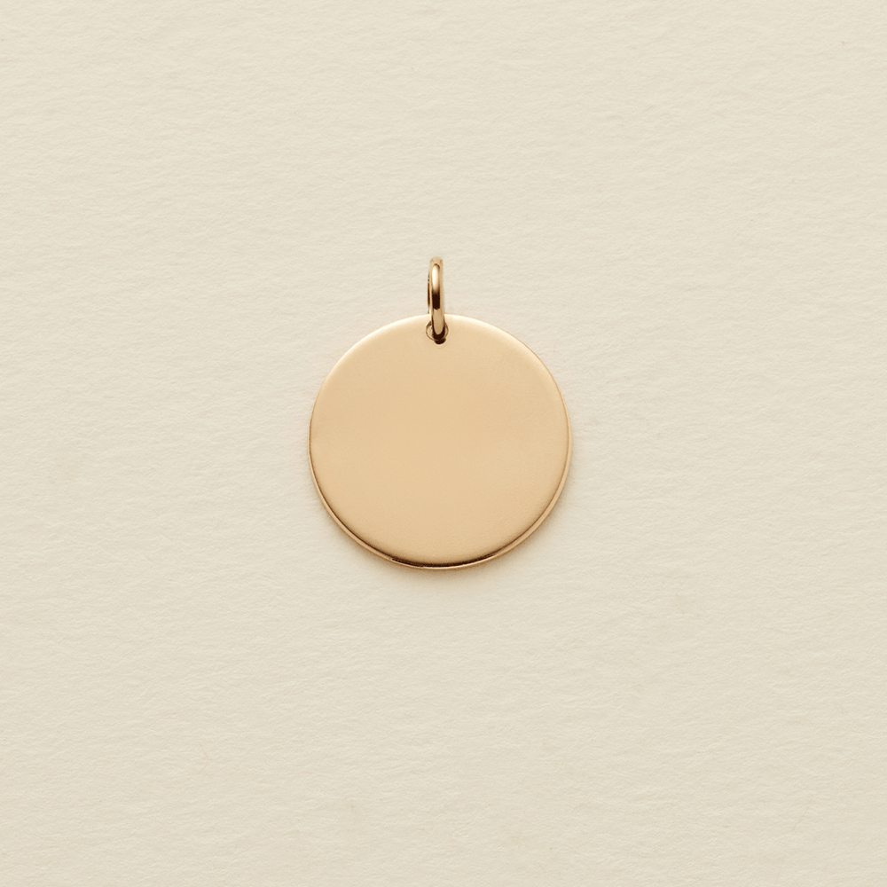 A flat round gold pendant with a jump ring on the top, shown on a textured light beige surface.