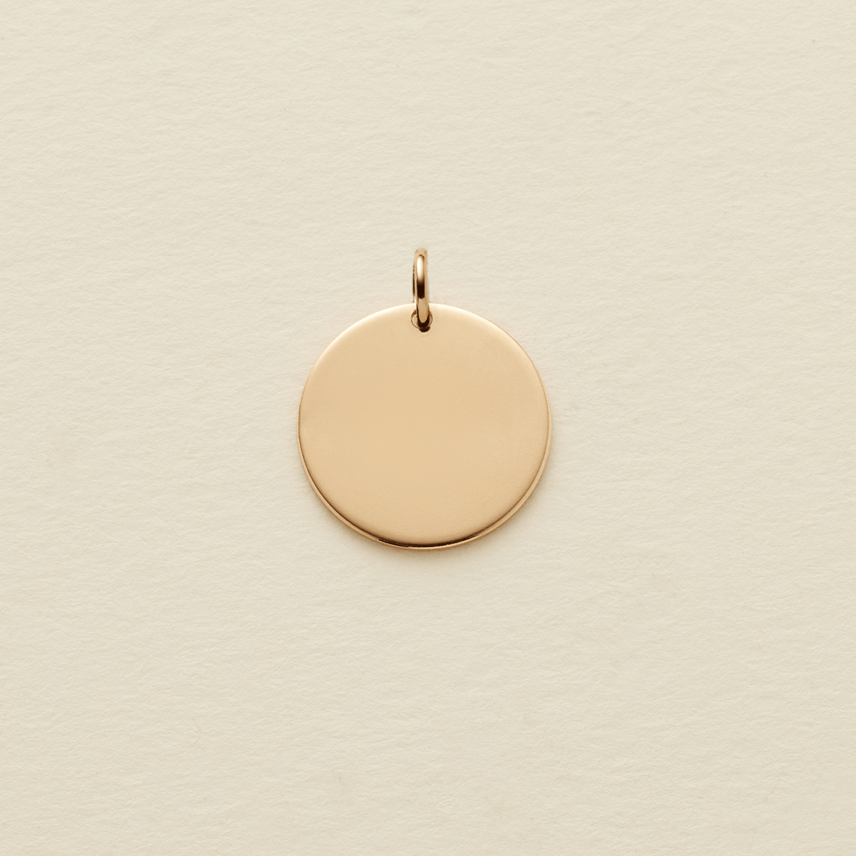 A flat round gold pendant with a jump ring on the top, shown on a textured light beige surface.