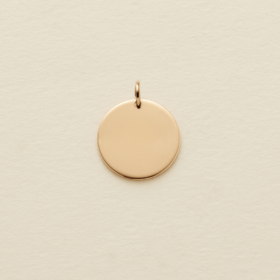 A flat round gold pendant with a jump ring on the top, shown on a textured light beige surface.