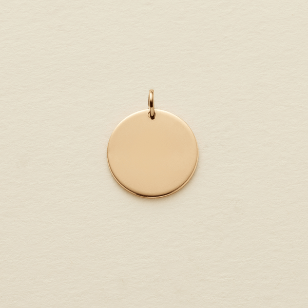 A flat round gold pendant with a jump ring on the top, shown on a textured light beige surface.