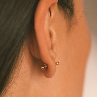 A close-up of a person's ear from the side, showing a gold bead stud earring with a round ball backing.