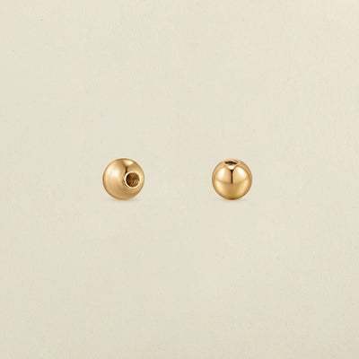A pair of polished round gold screw-on earring backings, shown on a solid light beige surface.