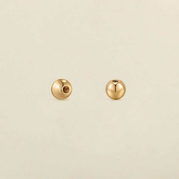 A pair of polished round gold screw-on earring backings, shown on a solid light beige surface.